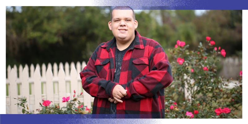 A person stands outdoors in a garden beside a white picket fence and flowering rose bushes. The person is wearing a red and black checkered jacket and is facing the camera. The image represents people with learning disabilities and highlights the importance of equitable access to healthcare and cancer care.