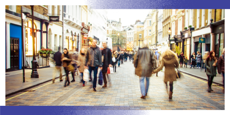 A busy pedestrian shopping street with people walking along a wide paved road lined with shops and buildings. The image is slightly blurred to convey movement and activity, showing members of the public moving through an urban environment. The scene illustrates community-based healthcare and public health settings outside of hospitals.