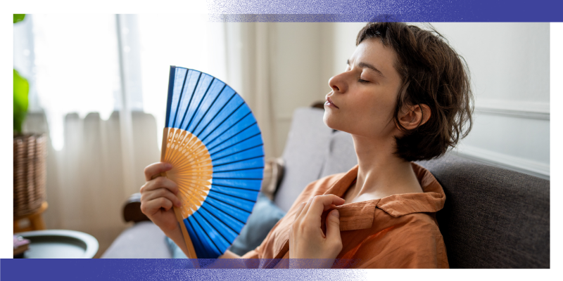 A person sits indoors beside a window, holding a handheld fan while cooling themselves during hot weather. The image illustrates the health impacts of heat and heatwaves, particularly for people living with long-term health conditions, and highlights the importance of protecting vulnerable individuals during periods of extreme temperature.