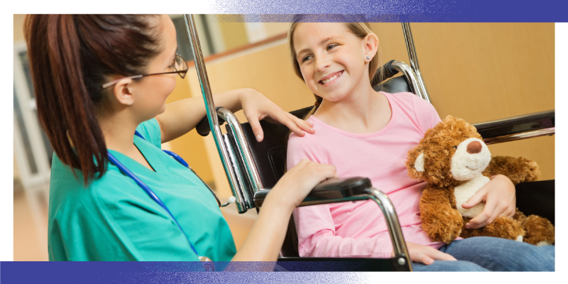 A healthcare professional in scrubs sits beside a child in a wheelchair during a hospital consultation. The child is holding a teddy bear while talking with the healthcare professional. The image illustrates paediatric healthcare, communication between patients and healthcare professionals, and support for children and families during medical care.