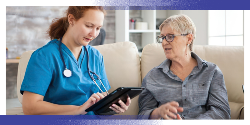 A healthcare professional wearing blue scrubs sits beside a patient on a sofa during a consultation. The healthcare professional is using a tablet device to record information while the patient gestures during the discussion. The image illustrates communication between patients and healthcare professionals and shared decision-making in a healthcare setting.