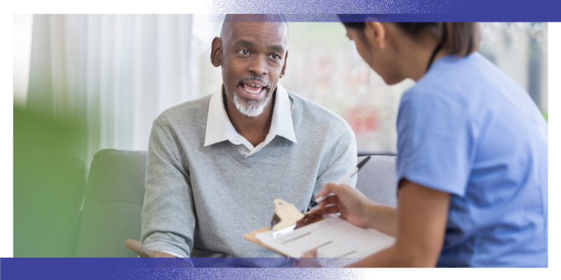 A healthcare professional sits with a patient during a consultation. The patient is seated on a sofa while the healthcare professional holds a clipboard and takes notes. The image illustrates a discussion about health, treatment or ongoing care in a community or outpatient healthcare setting.
